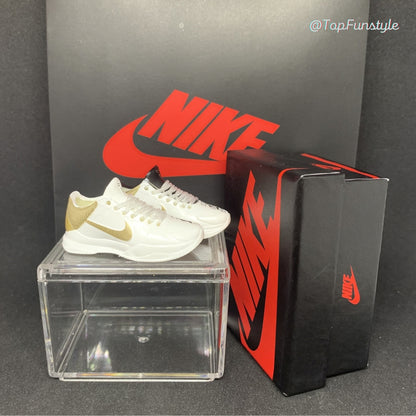 Mini sneakers Kobe 5 Big Stage Parade blanc et or posées sur la vitrine, boîte miniature à droite