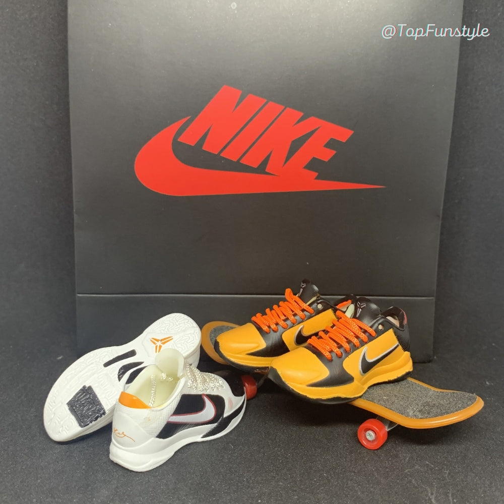 Les deux coloris Kobe 5 côte à côte : Bruce Lee jaune et noir sur un mini skateboard, Alternate blanc, noir et rouge